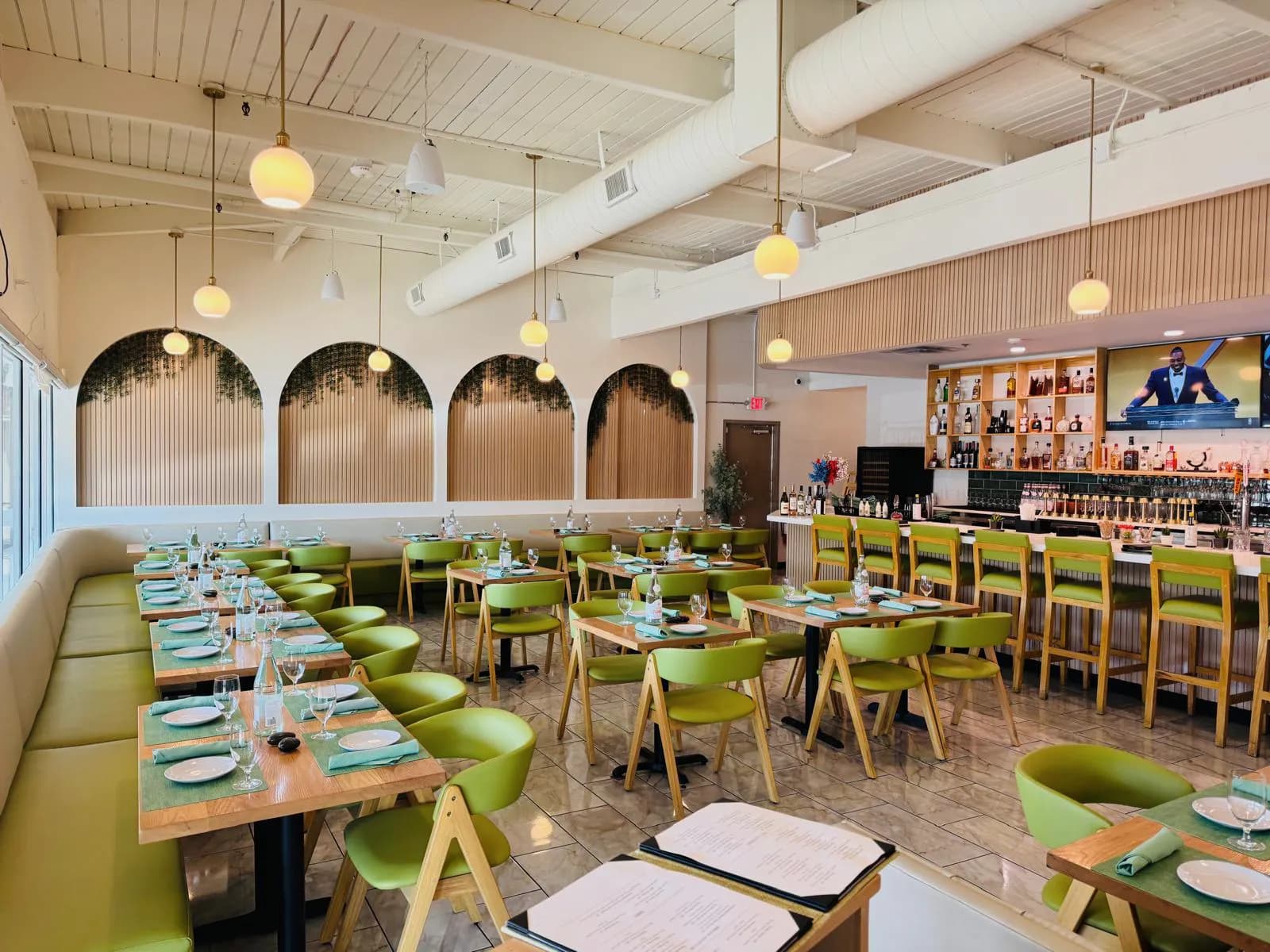 Wide Namak dining room with set tables, green seating, arched panels, and the bar beyond.